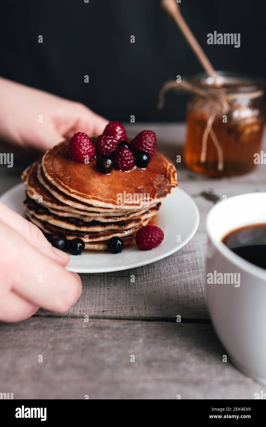 Hände halten weißen Teller mit Pfannkuchen, Beeren, Honig, Kaffeetasse auf Holztisch, Glas und Löffel. Hochwertige Fotos Stockfoto