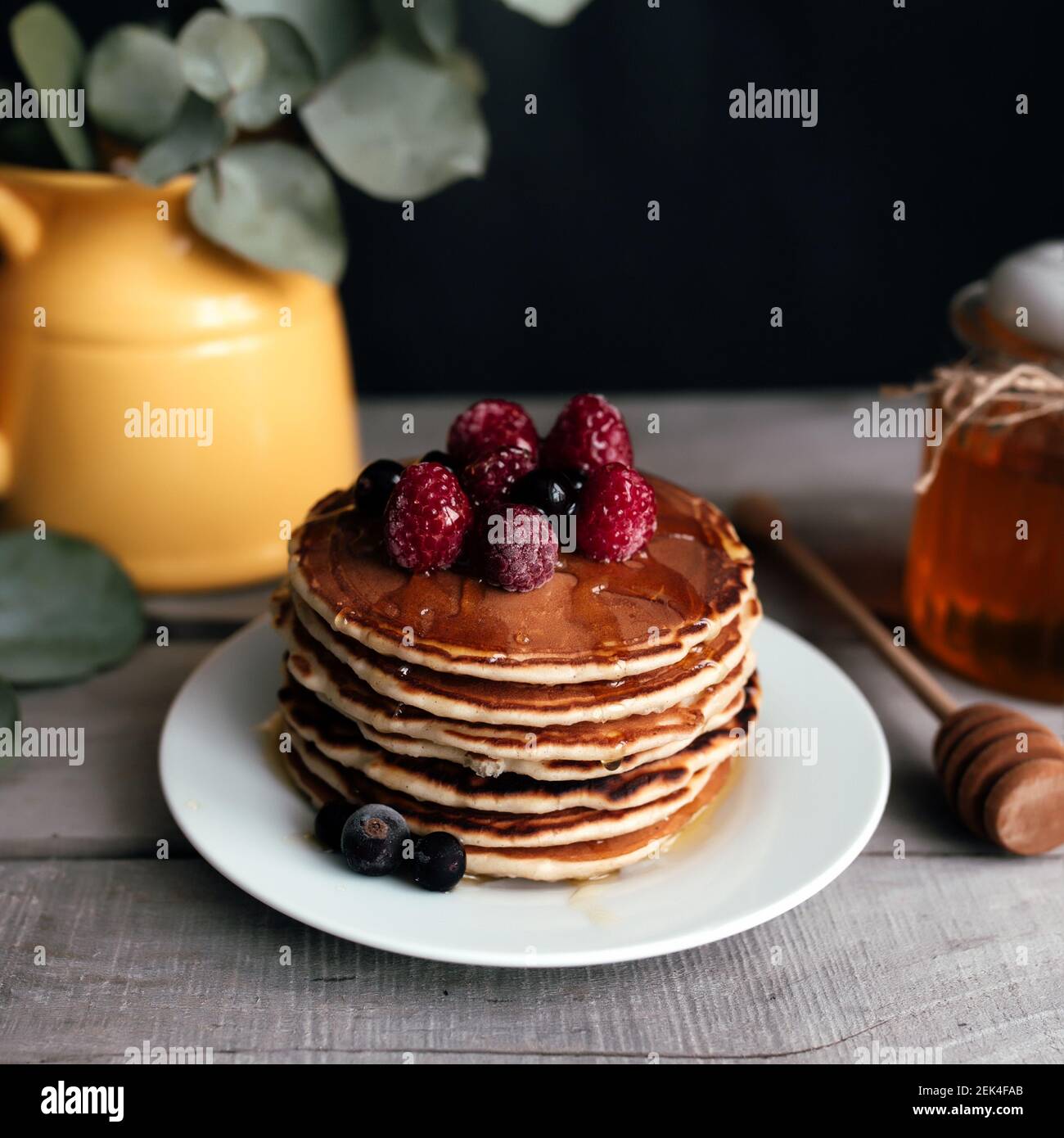 Saftige Pfannkuchen mit Beeren und Honig auf einem weißen Teller, Löffel, Glas, Holztisch, gelbe Vase mit Eukalyptus. Hochwertige Fotos Stockfoto