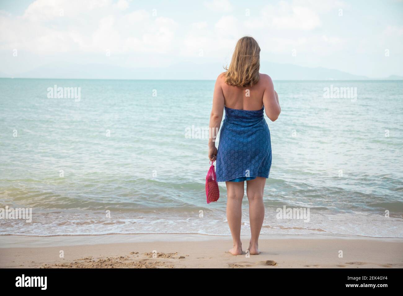 Eine Frau hält in der Hand eine Mesh-Einkaufstasche und steht am Rande des Meeres an einem Sandstrand. Ökologie-Konzept. Bewusster Konsum Stockfoto