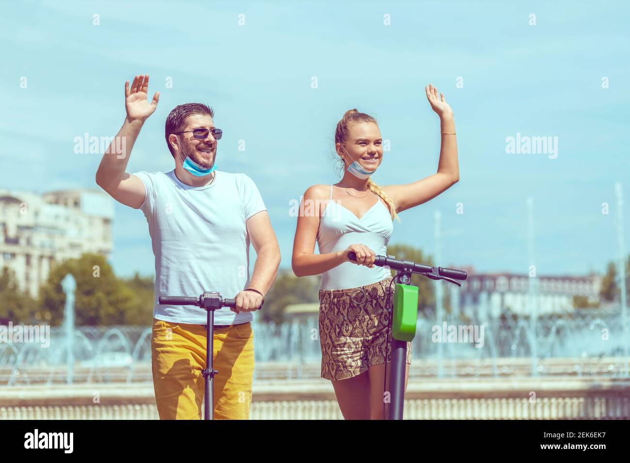 Glückliches Paar mit offener Gesichtsmaske im Urlaub Spaß haben Fahren Elektroroller im Stadtzentrum, während Freunde zu grüßen Stockfoto