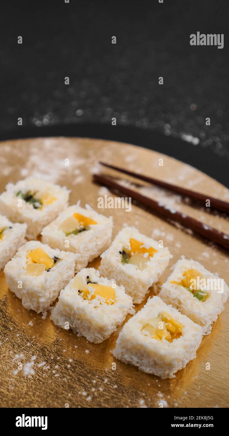 Sushi-Lieferung. Süße Brötchen aus Reis, Ananas, Kiwi und Mango. Rollt auf einem goldenen und schwarzen Hintergrund. Holzstöcke für Sushi. Stockfoto