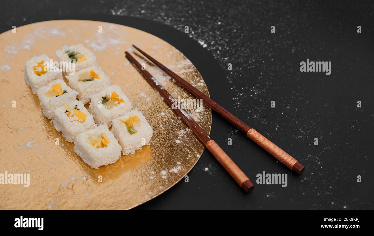 Sushi-Lieferung. Süße Brötchen aus Reis, Ananas, Kiwi und Mango. Rollt auf einem goldenen und schwarzen Hintergrund. Holzstöcke für Sushi. Stockfoto