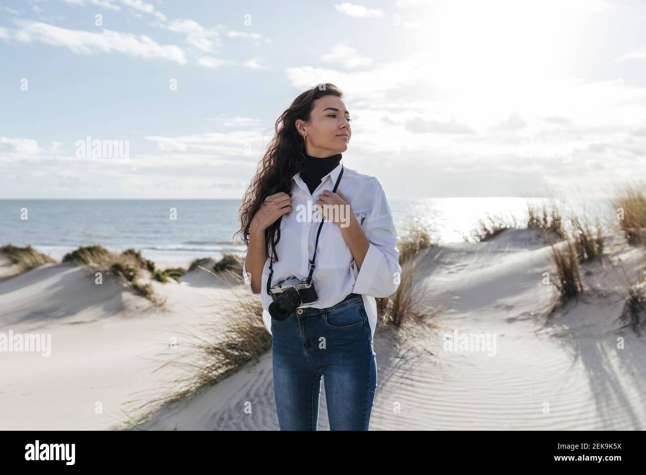 Junge Frau mit Vintage-Kamera steht auf Sanddünen gegen Himmel am Strand Stockfoto Junge Frau mit Vintage-Kamera steht auf Sanddünen gegen Himmel am Strand Stockfoto