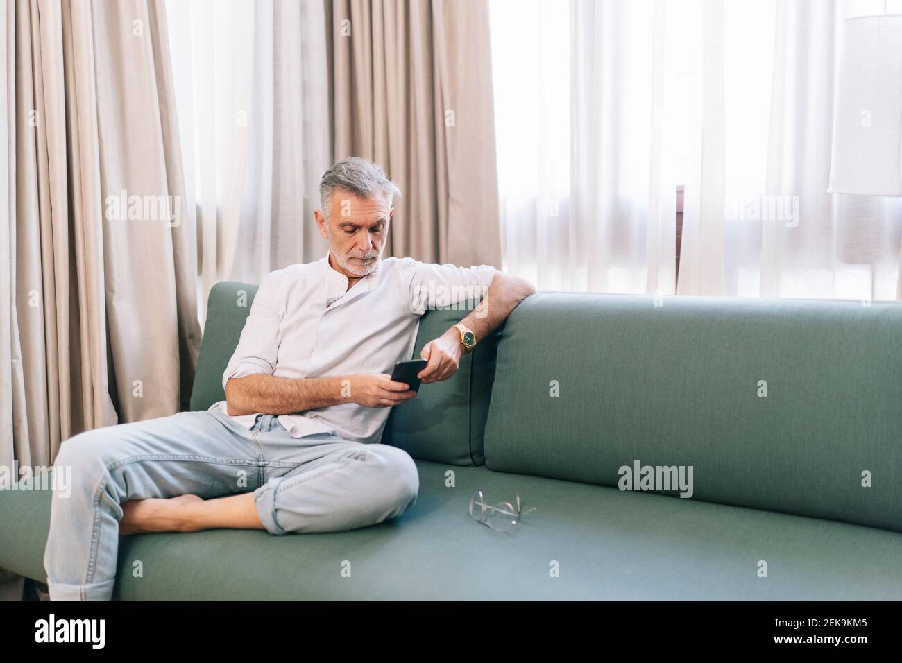 Mann, der Smartphone benutzt, während er auf dem Sofa gegen Vorhänge sitzt Im Hotelzimmer Stockfoto