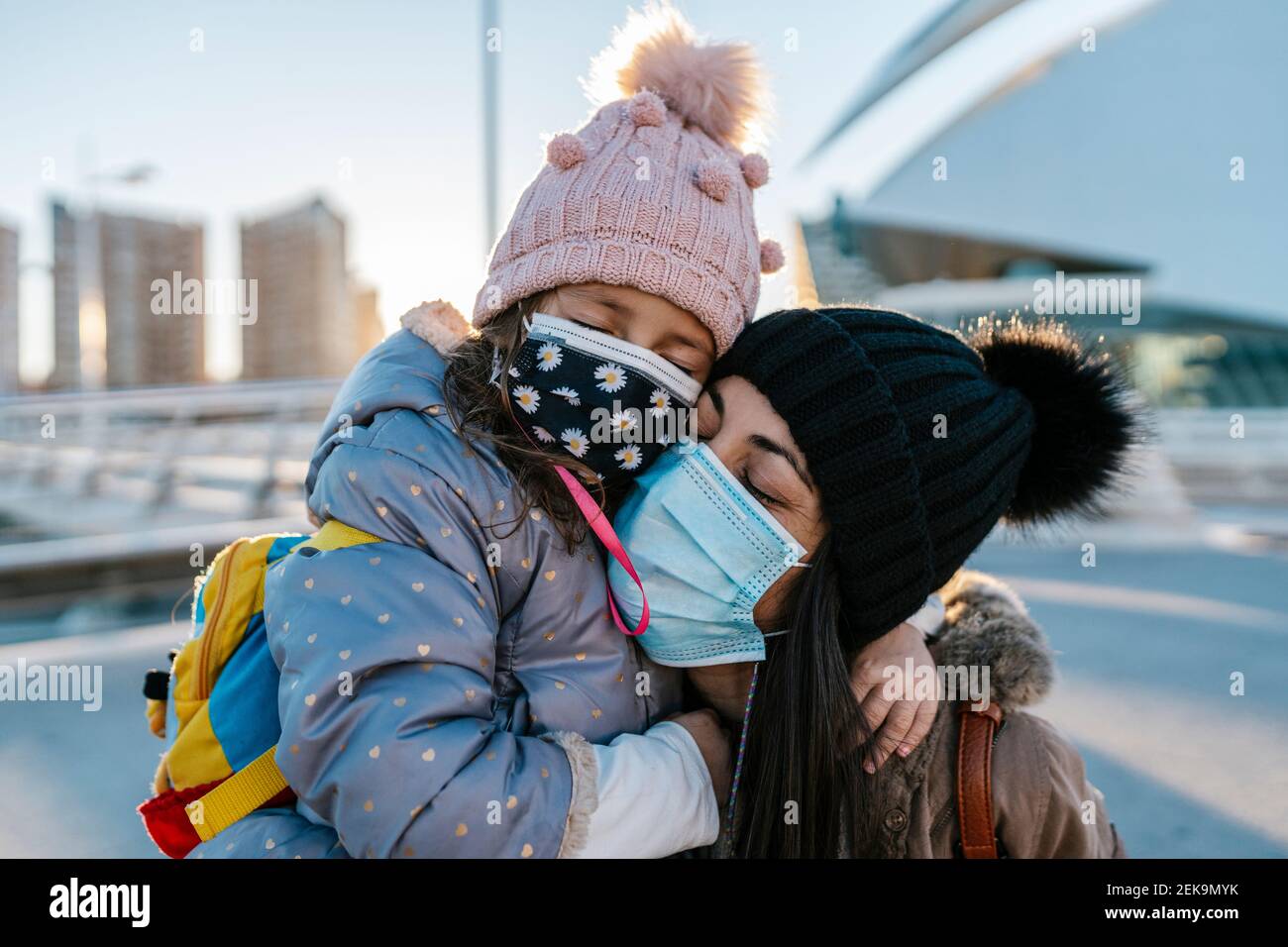 Mutter und Tochter tragen Strickmütze und Gesichtsmaske umarmt Im Freien stehen Stockfoto