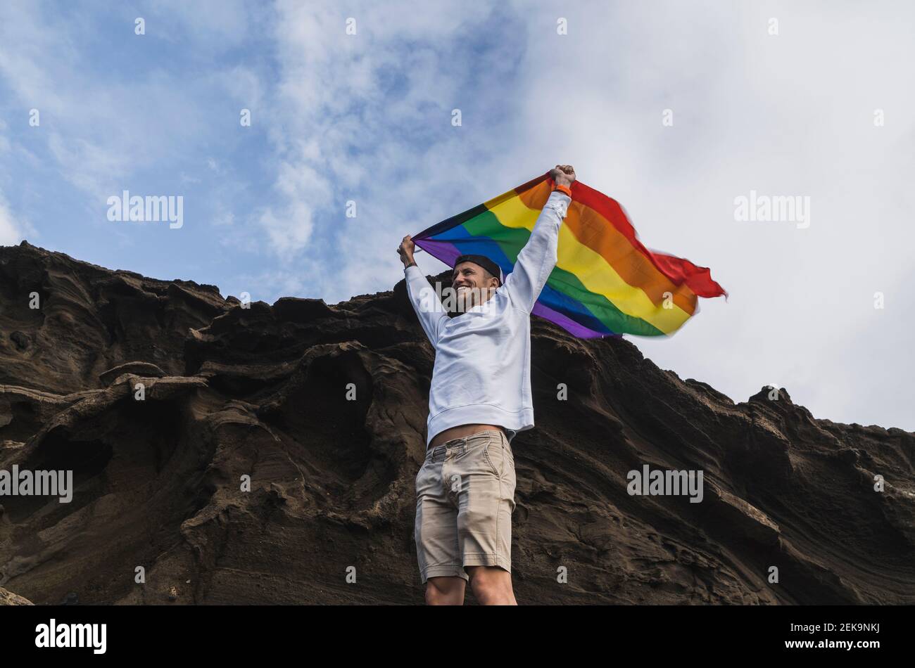 Lächelnder männlicher Tourist, der die LGBTQI-Flagge schwenkt, während er am Felsen in El golfo, Lanzarote, Spanien, steht Stockfoto