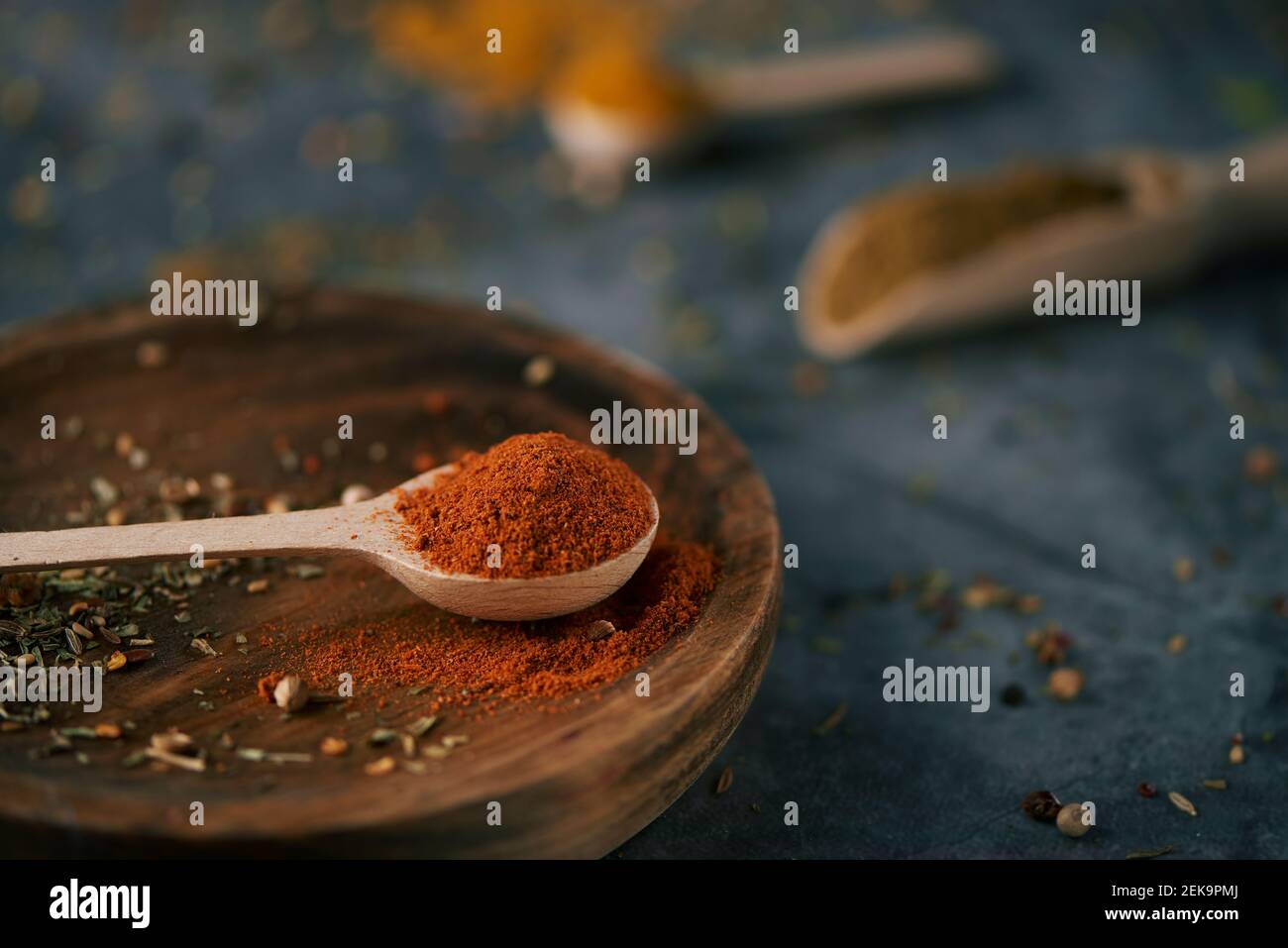 Nahaufnahme eines Holzlöffels voller rotem Currypulver, auf einem Holzteller, platziert auf dunkler Steinoberfläche und einigen anderen Holzlöffeln und -Kugeln mit Diffe Stockfoto