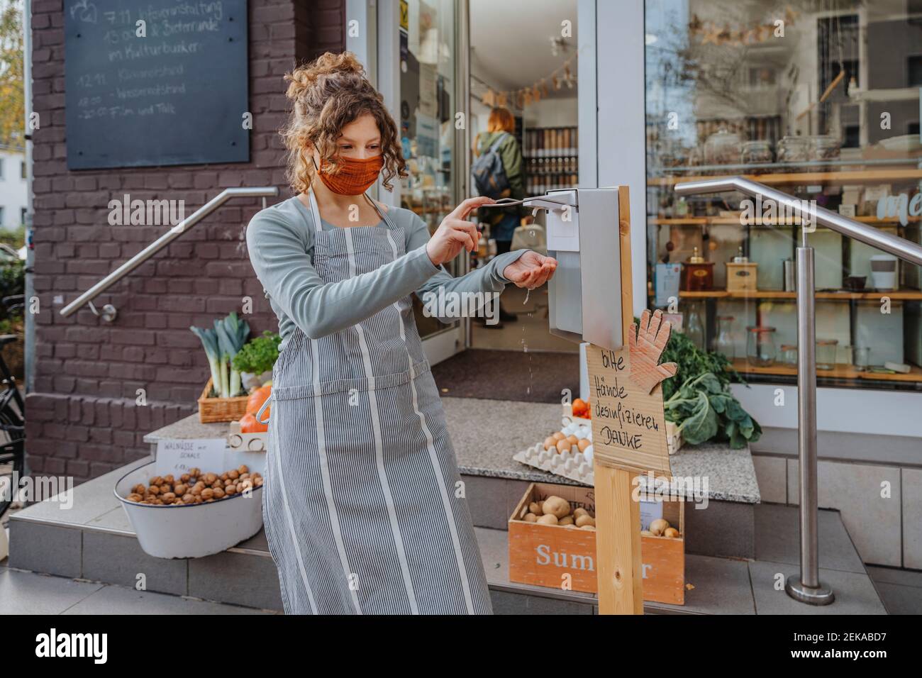 Weibliche Angestellte in der Schürze, die Händedesinfektionsmittel außerhalb des Bio-Ladens anwendet Stockfoto