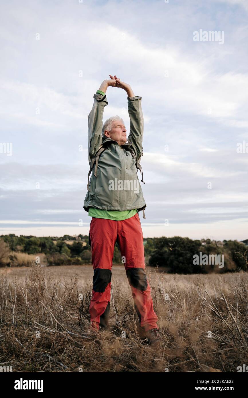 Älterer männlicher Wanderer, der die Arme streckt, während er auf dem landwirtschaftlichen Feld steht Auf dem Land ist der Himmel bewölkt Stockfoto