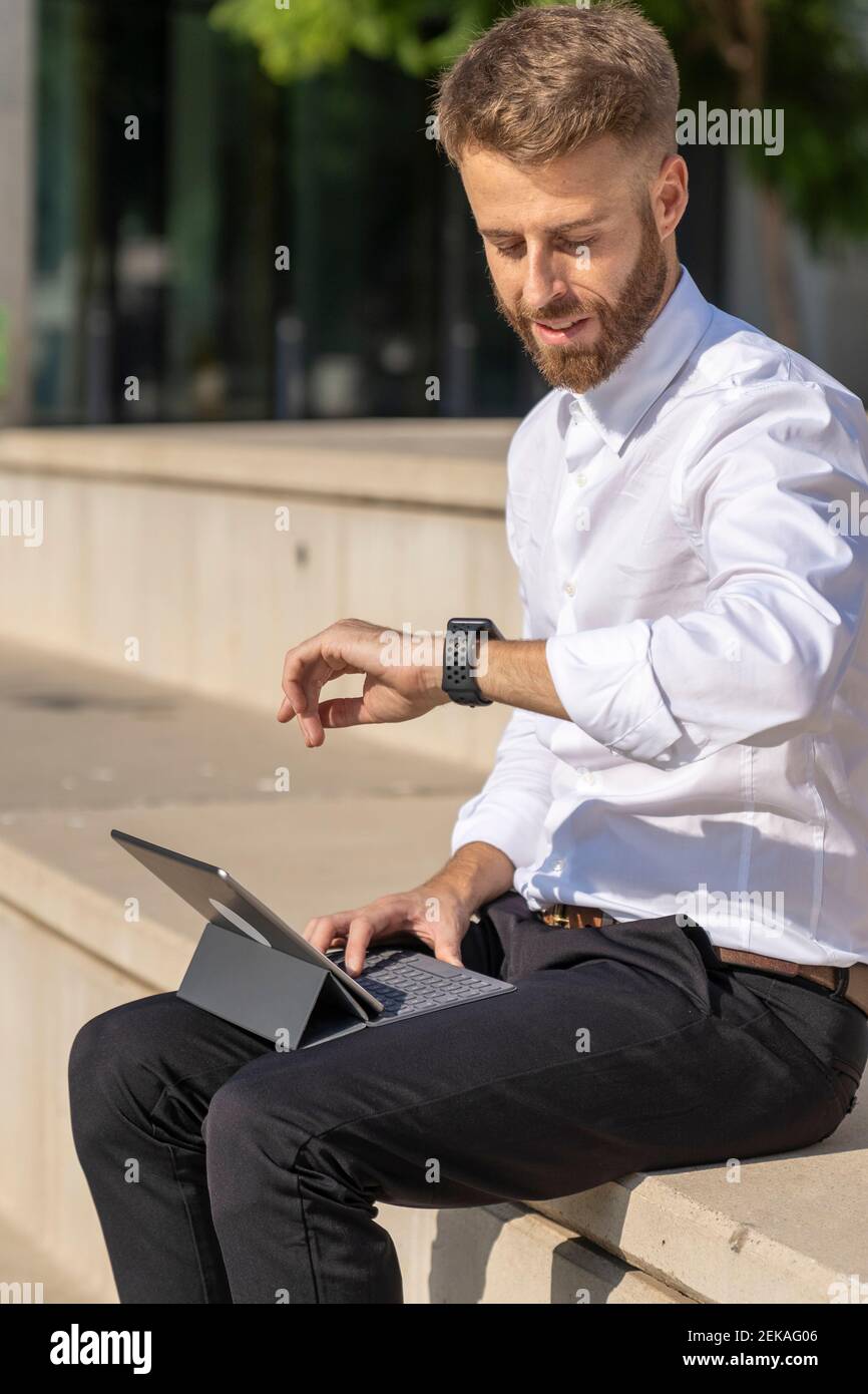 Geschäftsmann, der die Zeit auf der Uhr überprüft, während er mit einem digitalen Tablet sitzt Auf Stufen Stockfoto