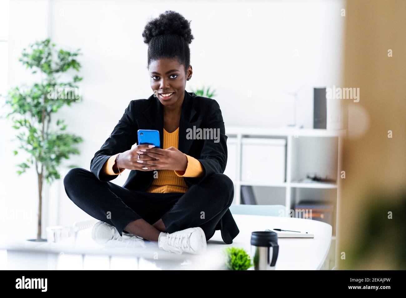 Lächelnd junge Afro weibliche professionelle Textnachrichten mit Handy Auf dem Schreibtisch sitzend mit gekreuzten Beinen Stockfoto