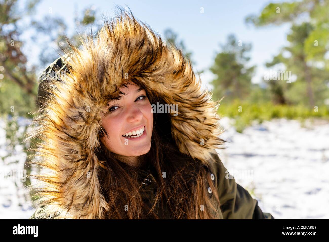 Lächelnde junge Frau trägt Fell Kapuzen Wintermantel starren während Im Wald stehen Stockfoto