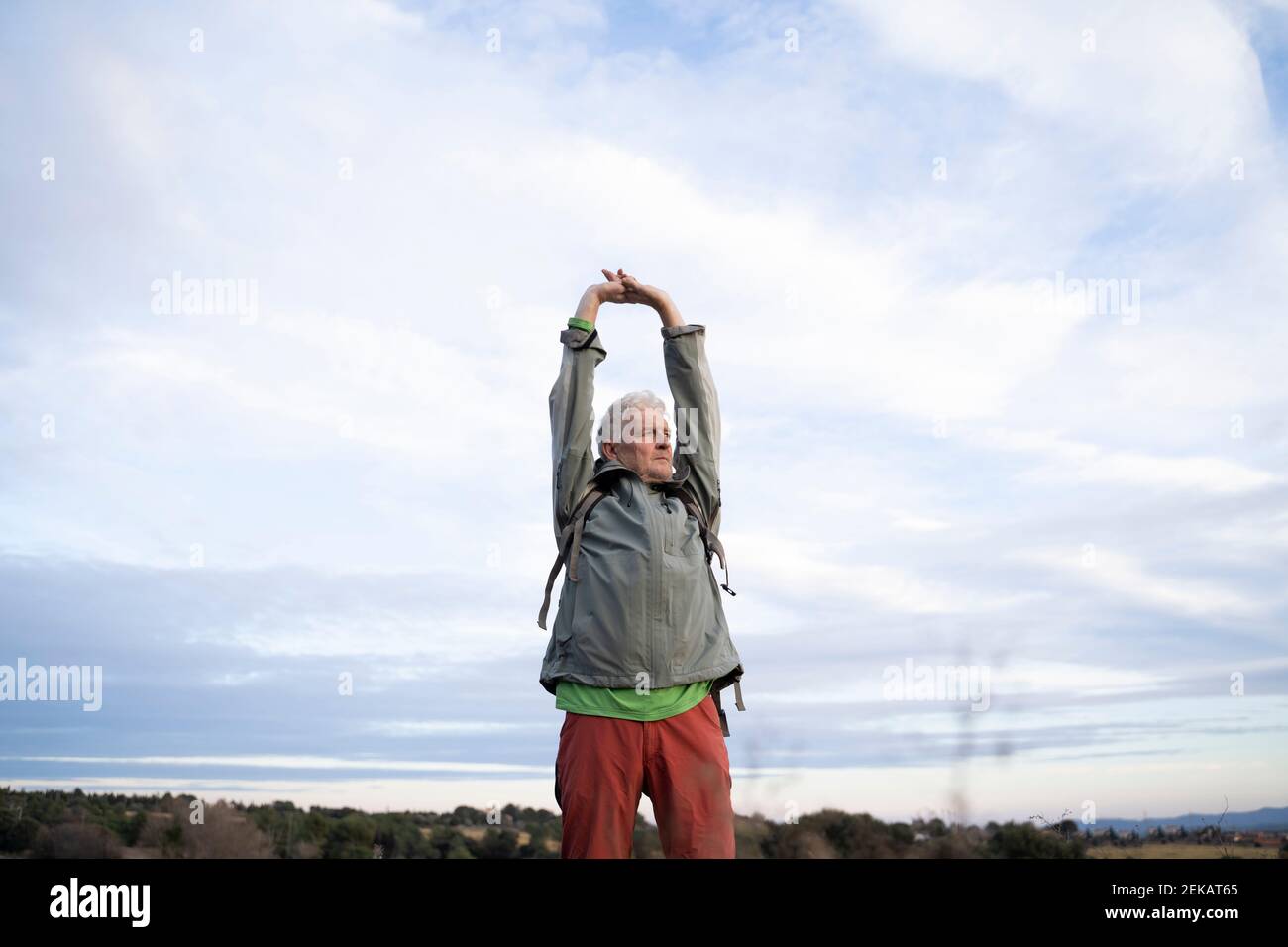 Älterer männlicher Wanderer, der die Arme streckt, während er gegen den bewölkten Himmel steht Auf dem Land Stockfoto