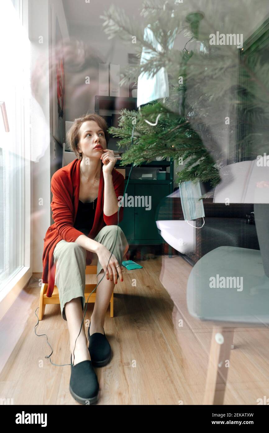 Frau sitzt am Weihnachtsbaum durch Glas zu Hause gesehen Während einer Pandemie Stockfoto