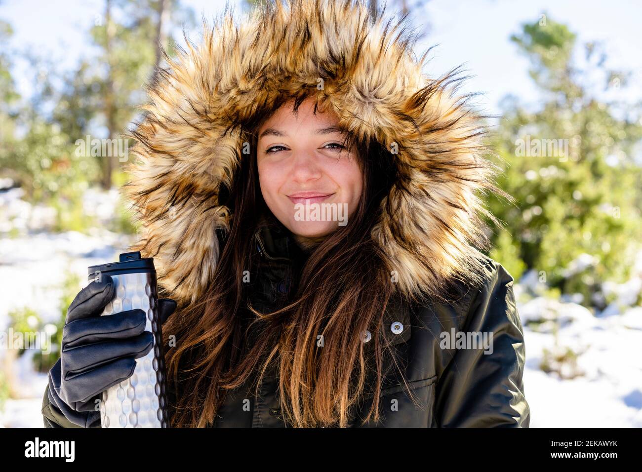 Lächelnde Frau trägt Pelz Kapuzen Wintermantel hält Flasche während Stehen am Wald Stockfoto