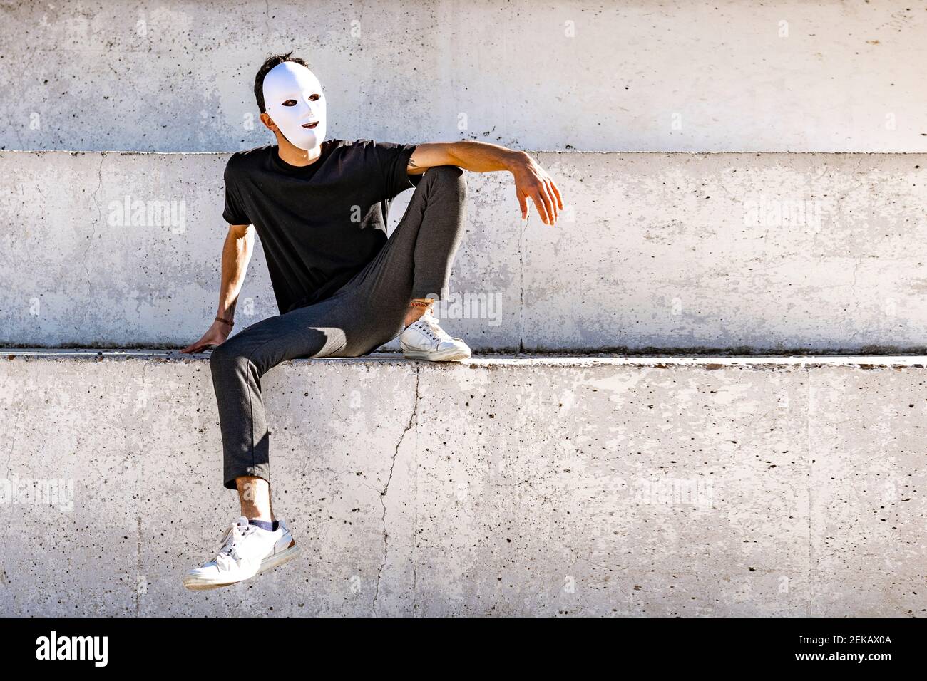 Junger Mann mit weißer Gesichtsmaske schaut weg, während Sitzen an der Wand Stockfoto