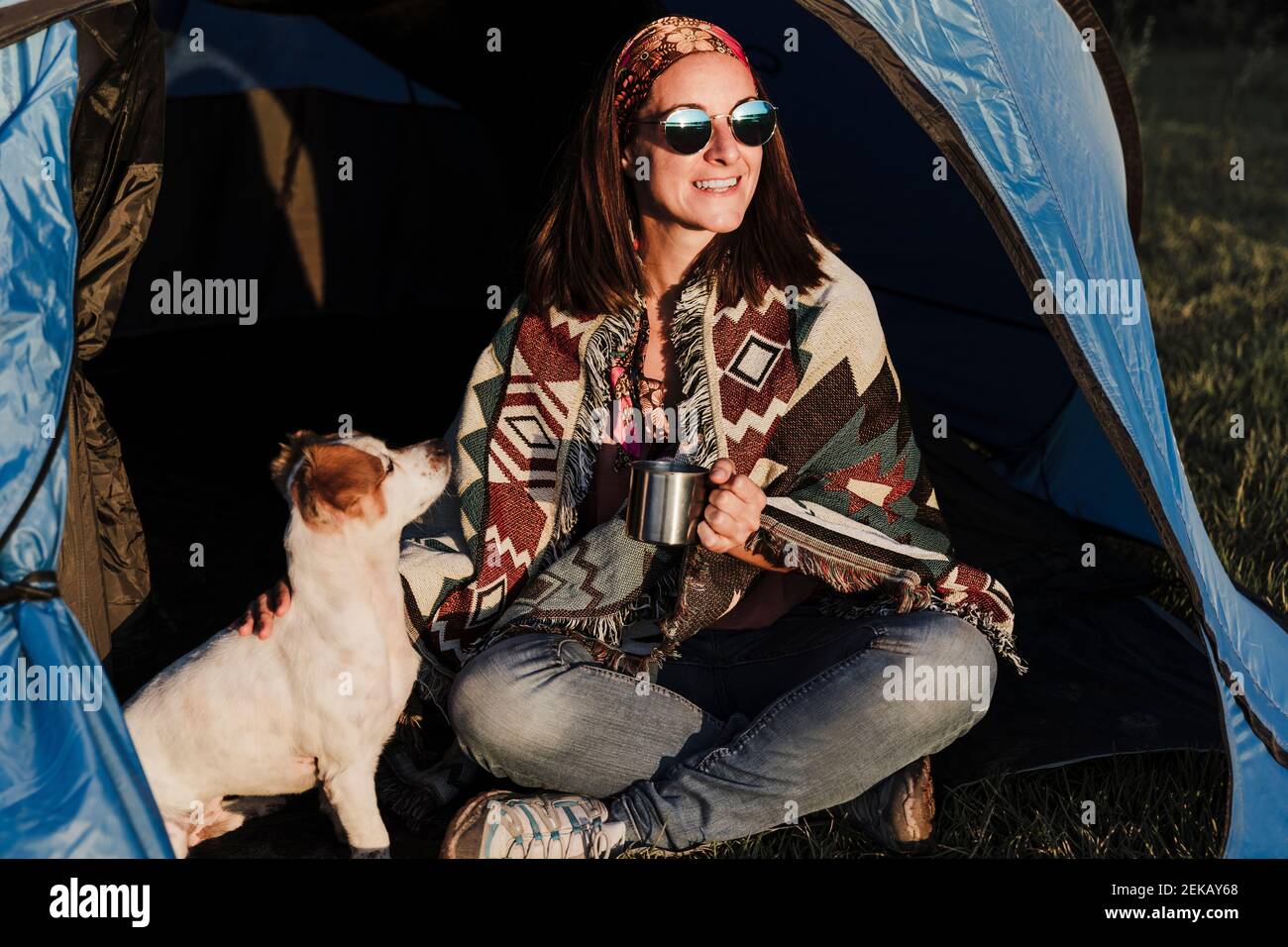 Glückliche Frau trägt eine Sonnenbrille und hält den Becher, während sie mit dem Hund sitzt Im Zelt Stockfoto