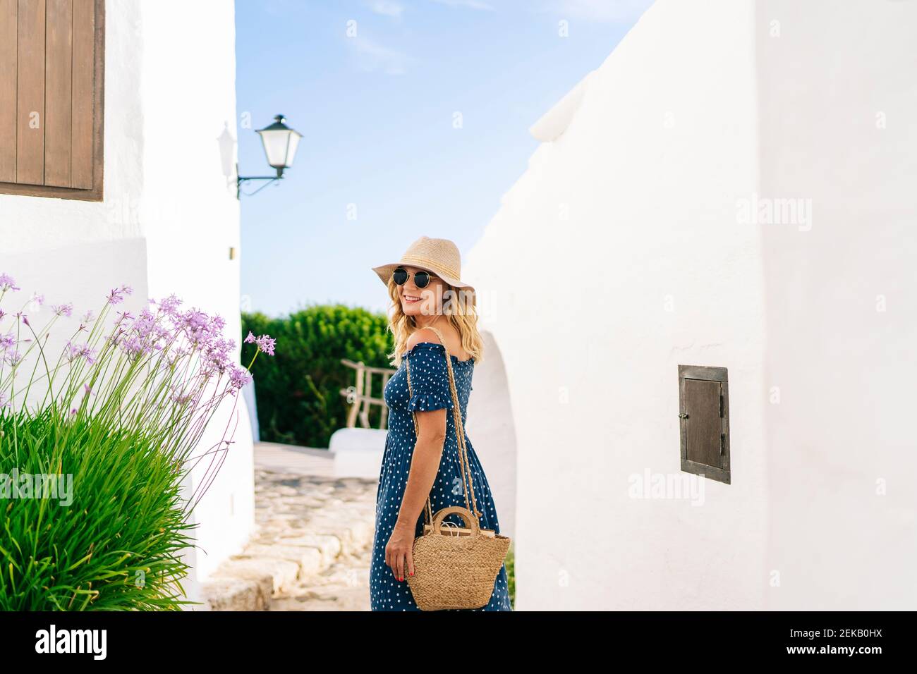 Weibliche Touristin in Sonnenbrillen und Hut, die über die Schulter schauen, während sie in Binibeca Dorf, Menorca, Spanien steht Stockfoto