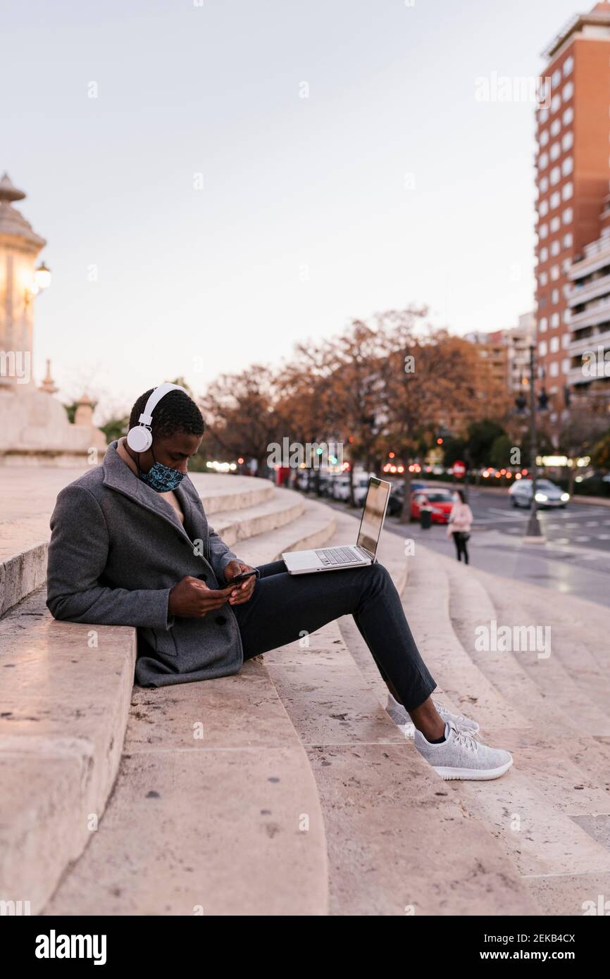 Mann trägt Gesichtsmaske mit Handy, während sitzend mit Laptop auf Stufen Stockfoto