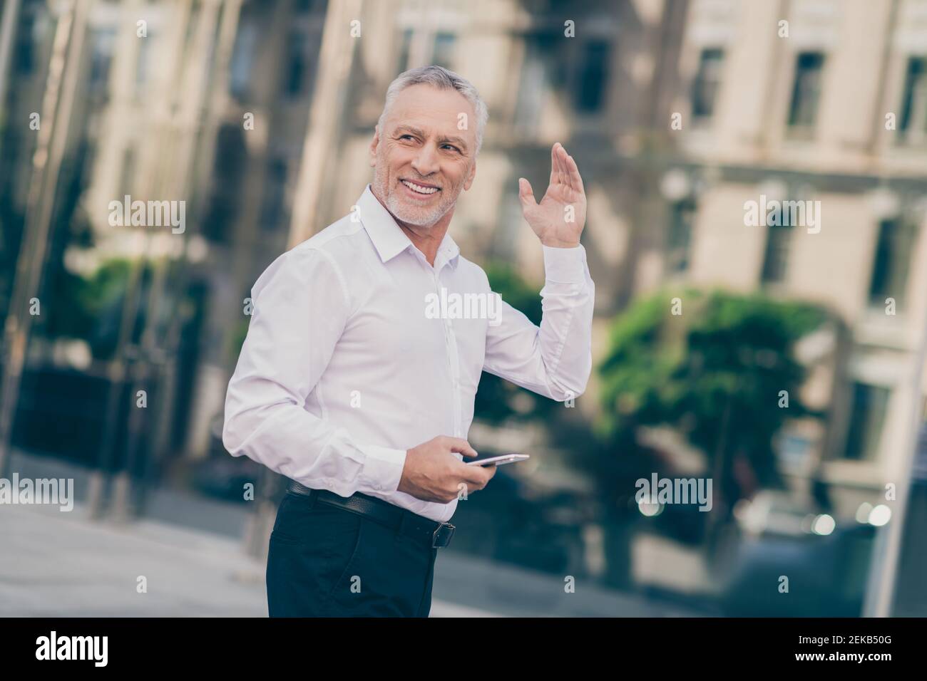 Profilfoto von optimistischen grauen Haaren alten Business-Mann Welle Telefon in der Hand tragen weißes Hemd im Freien in der Nähe des Arbeitszentrums Stockfoto