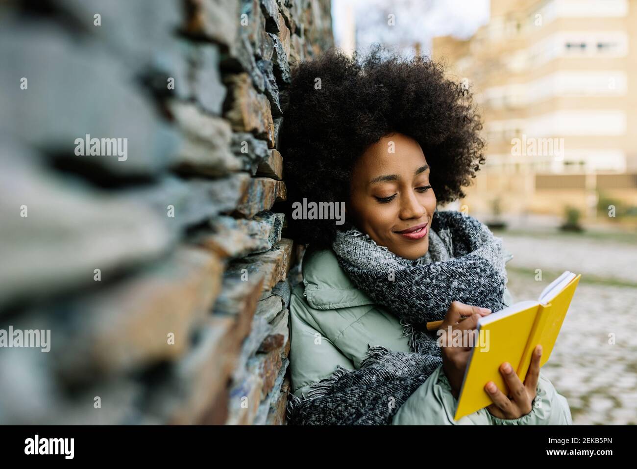 Lächelnde Afro-Frau, die in einem Buch schreibt, während sie sich auf Stein stützt Wand im Winter Stockfoto