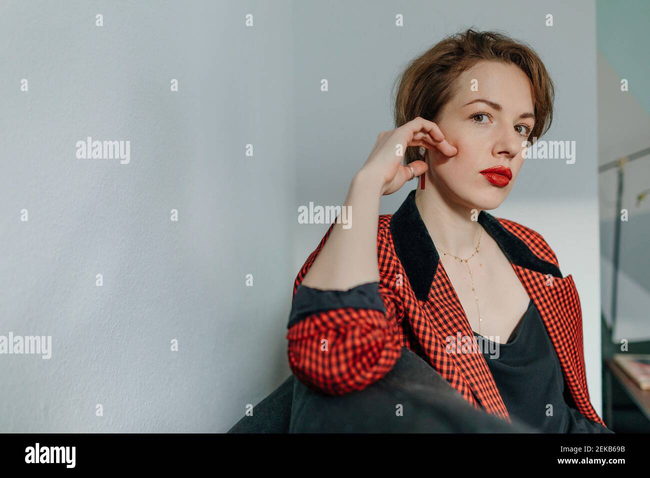 Selbstbewusste junge Frau mit rot kariertem Blazer, der gegen Weiß sitzt Wand Stockfoto