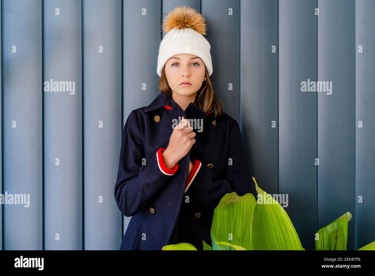 Ernsthafte Teenager-Mädchen tragen Wintermantel stehen von Zimmerpflanze gegen Wand zu Hause Stockfoto