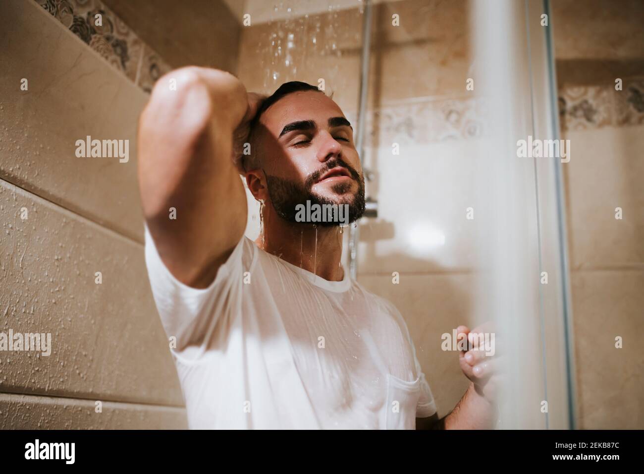 Junger Mann im T-Shirt mit der Hand im Haar unter der Dusche Im Badezimmer zu Hause Stockfoto