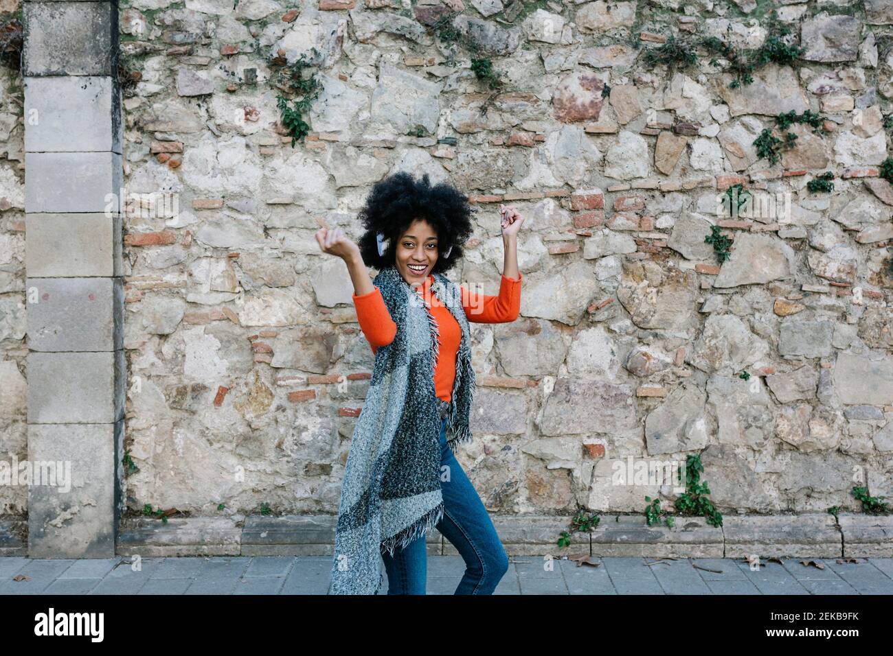 Fröhliche junge Frau tanzt, während Musik über Kopfhörer gegen Steinmauer Stockfoto