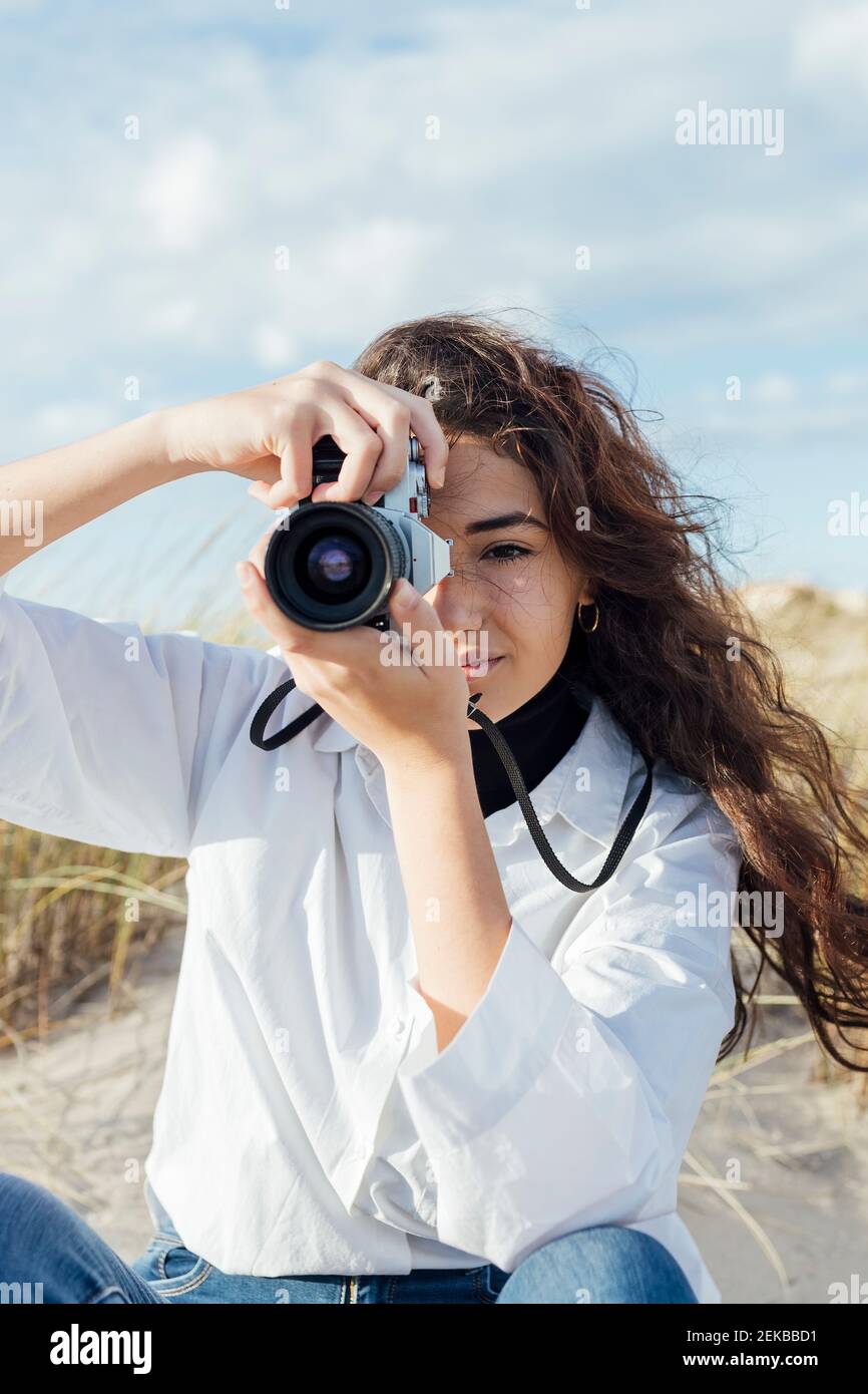 Junge Frau, die mit Vintage-Kamera fotografiert, während sie auf sitzt Sand gegen bewölkten Himmel Stockfoto Junge Frau, die mit Vintage-Kamera fotografiert, während sie auf sitzt Sand gegen bewölkten Himmel Stockfoto