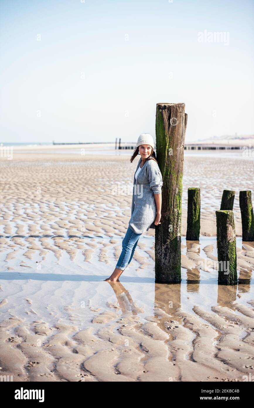 Junge Frau, die sich am Strand an einem Holzpfosten lehnte Tag Stockfoto