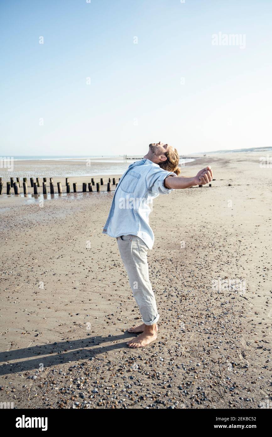 Junger Mann mit ausgestreckten Händen, der bei Sonnenschein am Strand steht Tag Stockfoto