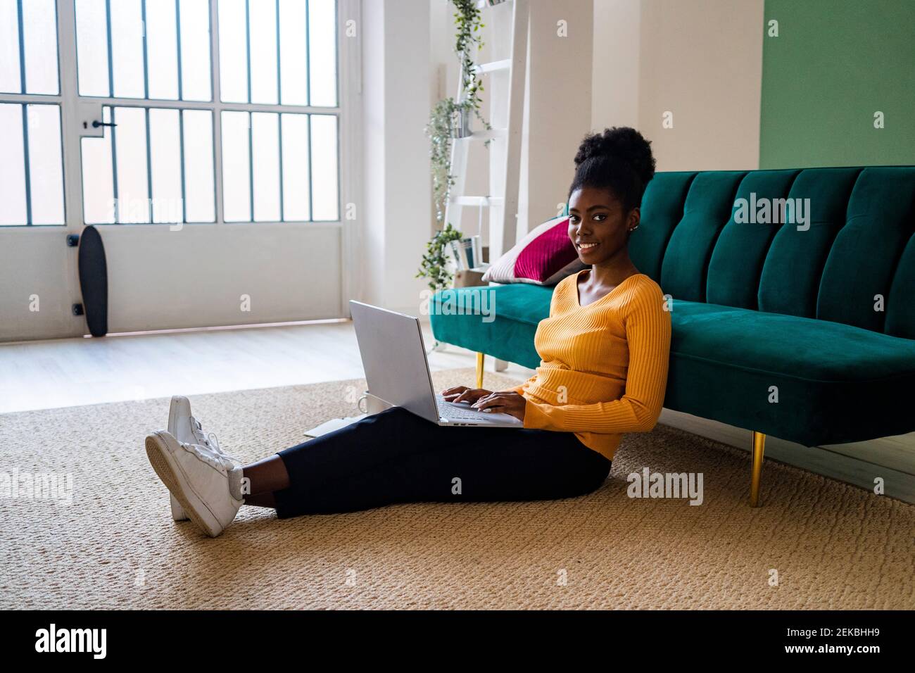 Lächelnde junge Afro-Frau, die mit Laptop auf dem Teppich sitzt Sofa im Wohnzimmer zu Hause Stockfoto