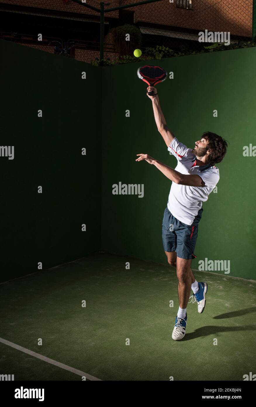 Männlicher Athlet, der den Ball bedient, während er nachts auf dem Sportplatz Padel-Tennis spielt Stockfoto