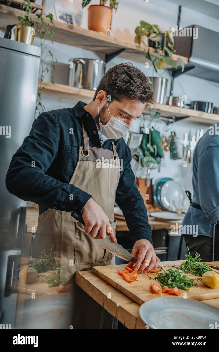 Mittlerer Erwachsener Koch trägt schützende Gesichtsmaske schneiden Gemüse, während Stehen mit Kollegen in der Küche Stockfoto