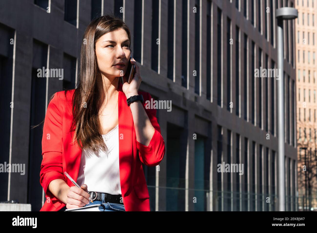 Weibliche Unternehmerin, die im Tagebuch schreibt, während sie auf dem Handy spricht Gegen Gebäude Stockfoto