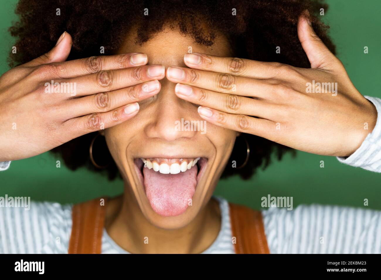 Fröhliche junge Frau, die die Zunge herausstreckt, während sie die Augen davor bedeckt Grüne Wand Stockfoto