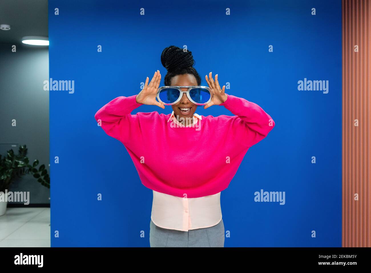 Fröhliche kreative weibliche Profi trägt übergroße Sonnenbrille an der blauen Wand Am Arbeitsplatz Stockfoto