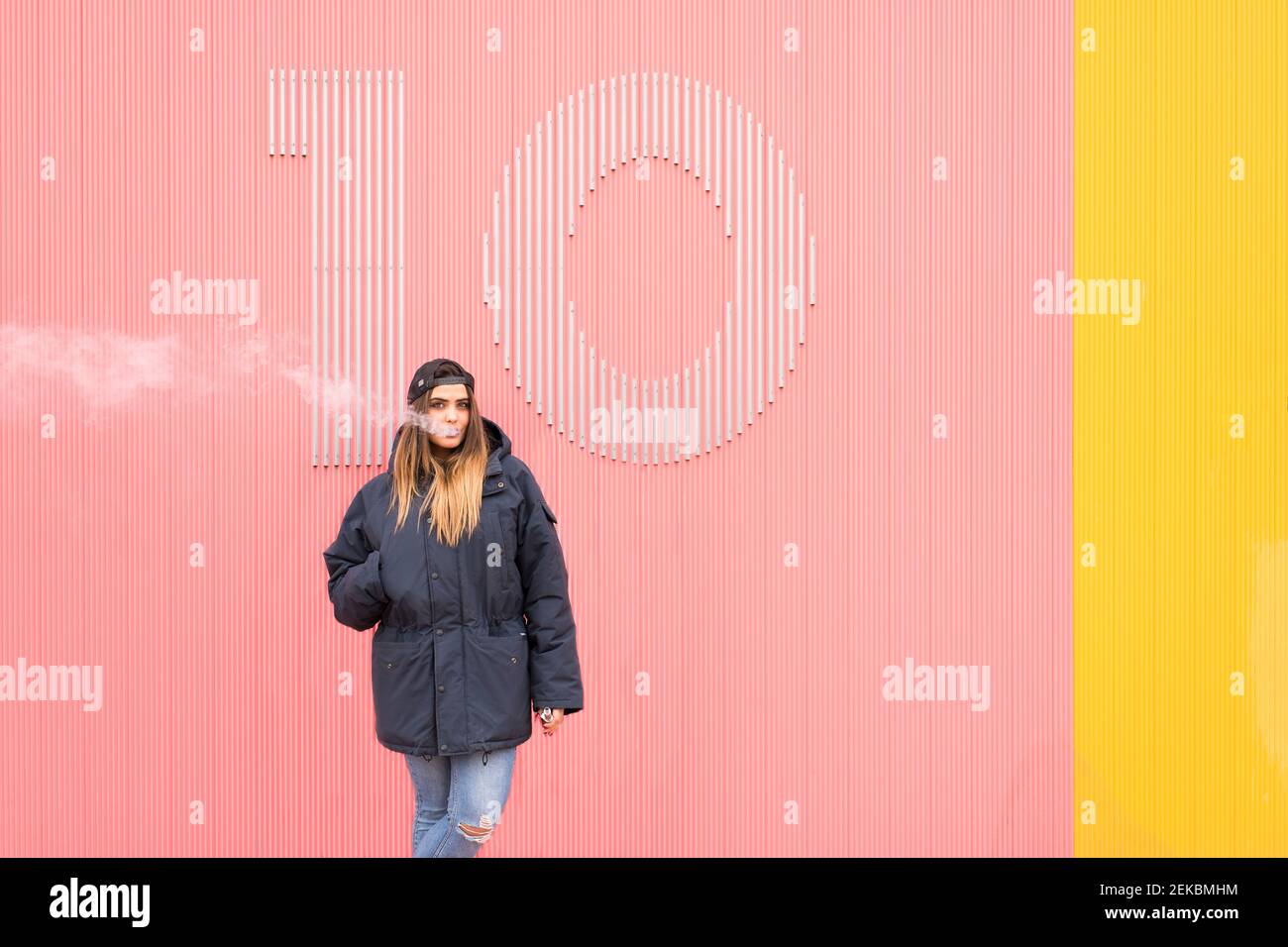 Junge Frau in Jacke atmet Rauch an rosa und gelben Wänden aus Stockfoto