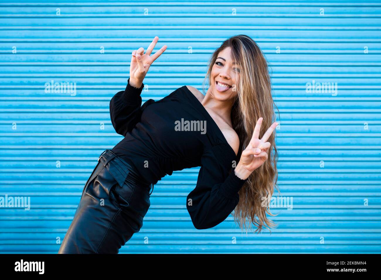 Fröhliche Frau, die die Zunge herausstreckt und Friedenszeichen dagegen zeigt Blaue Wand Stockfoto