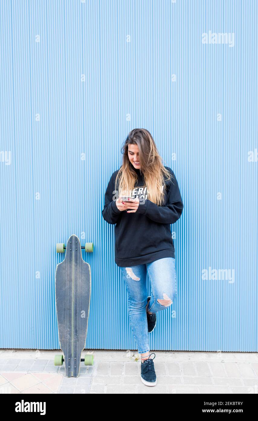 Junge Frau mit Skateboard und Smartphone, während sie auf einem Bein gegen die blaue Wand steht Stockfoto