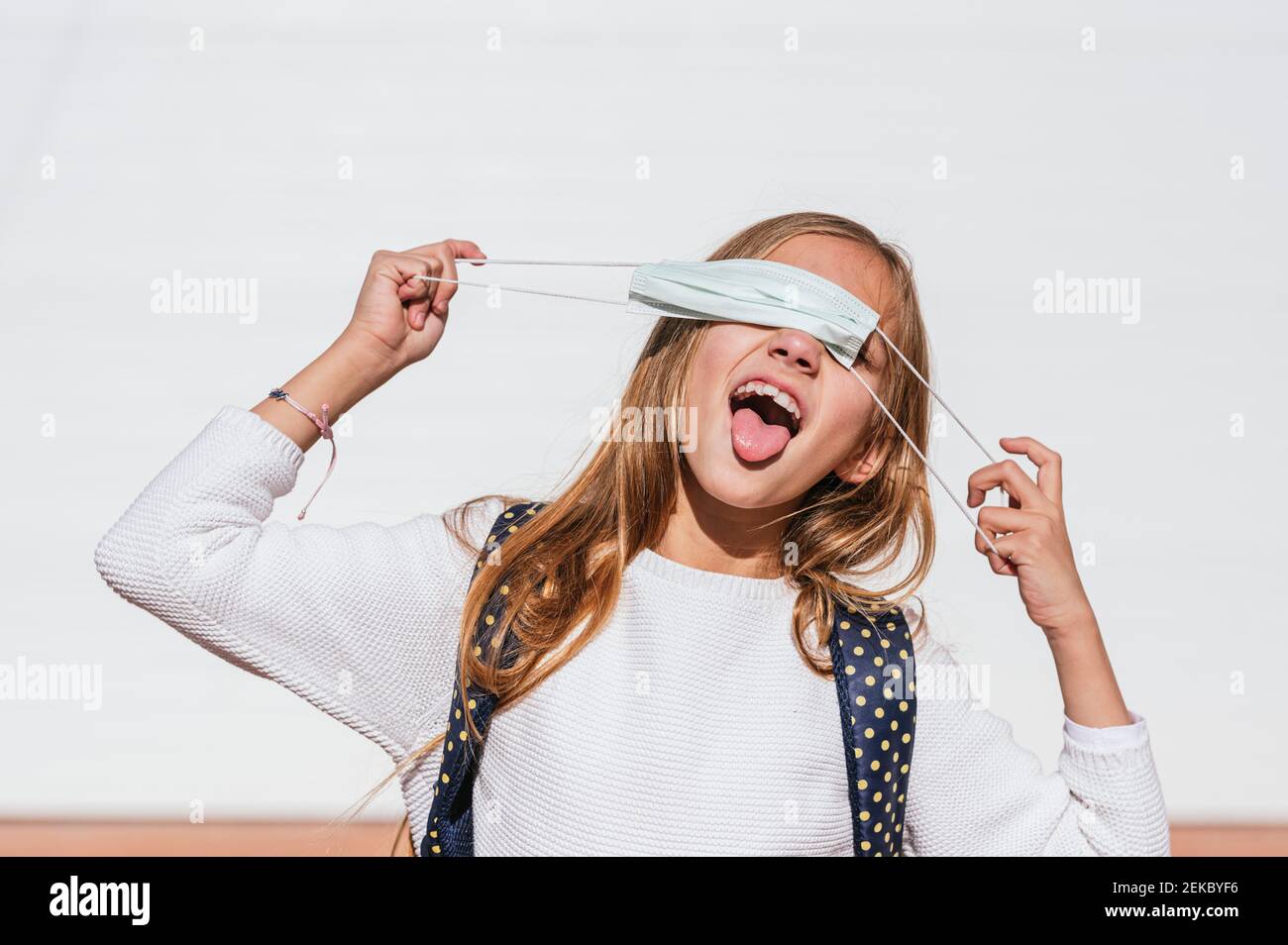 Verspielte Mädchen Abdeckung Auge mit schützenden Gesichtsmaske beim Kleben Die Zunge gegen die Wand während der Pandemie Stockfoto