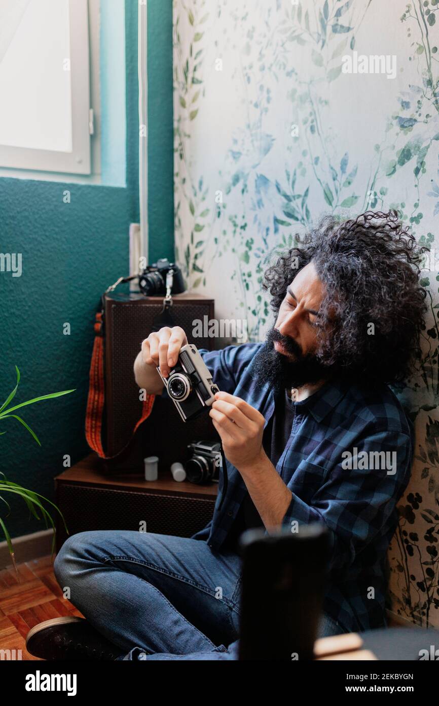 Hipster Mann hält Kamera, während er mit gekreuzten Beinen gegen die Wand sitzt Zu Hause Stockfoto