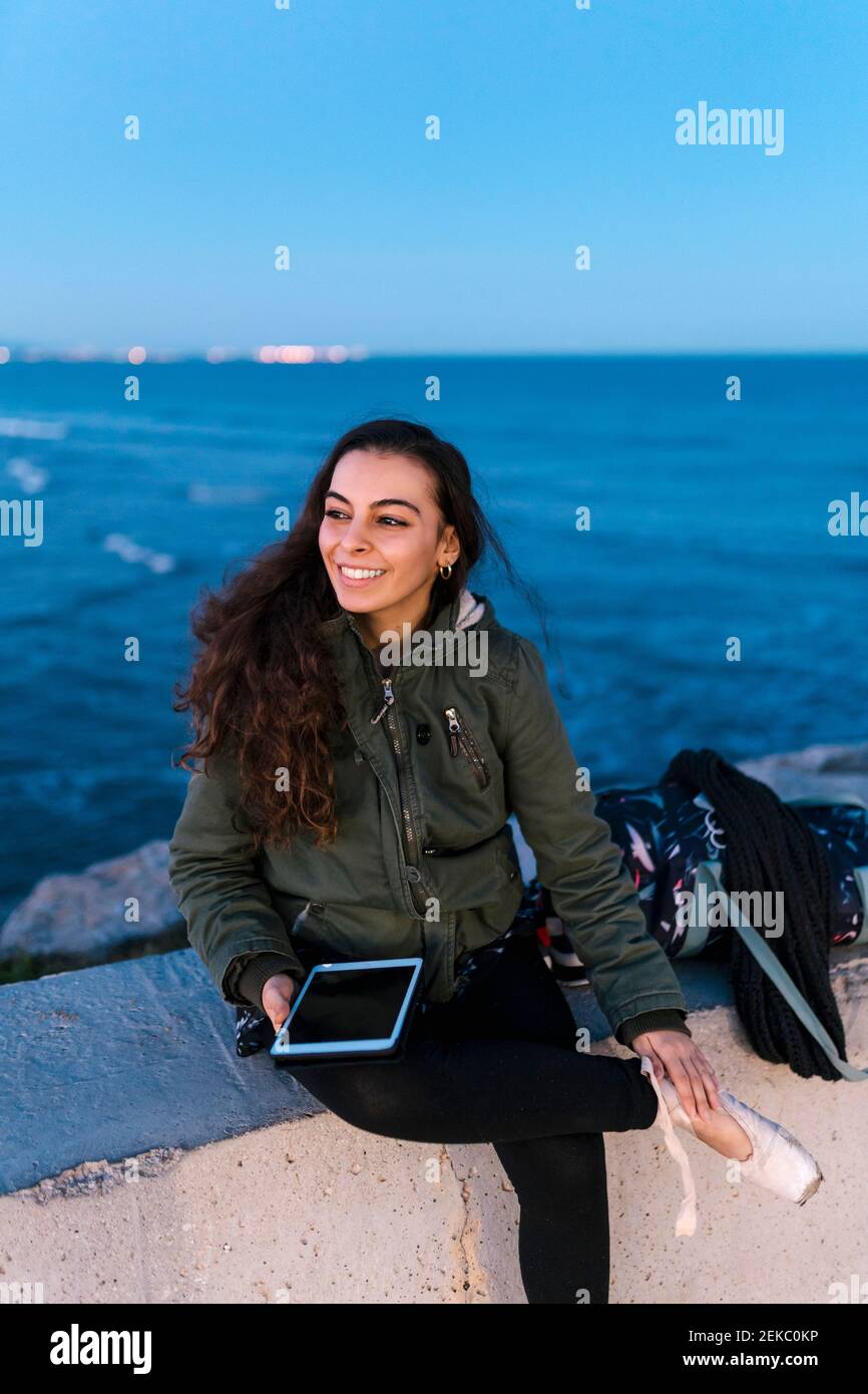 Schöne Frau mit digitalen Tablet sitzt auf Stützwand an Strand in der Dämmerung gegen Himmel Stockfoto