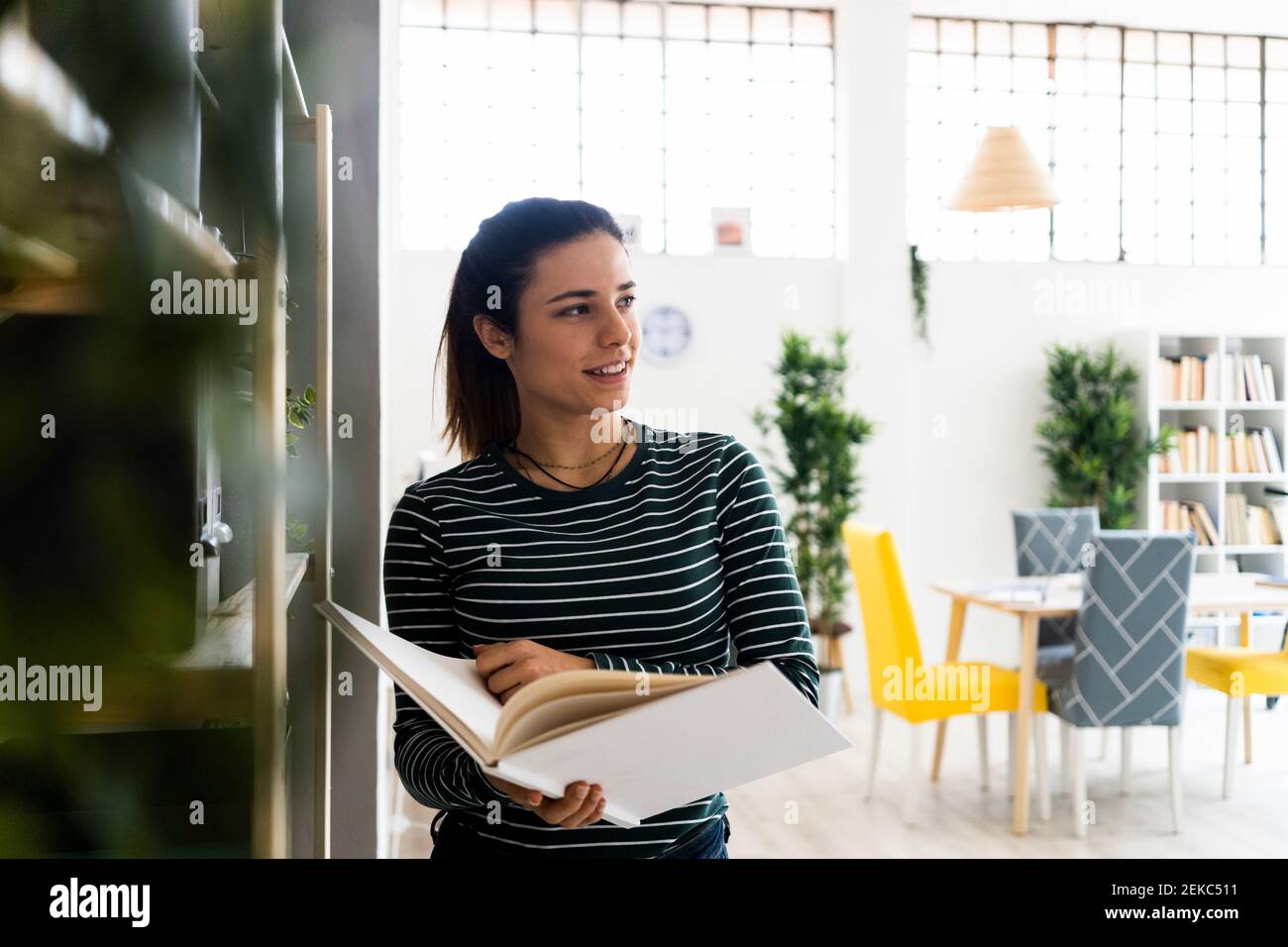 Lächelnde Unternehmerin hält Buch, während Tag träumen bei der Arbeit Ort Stockfoto