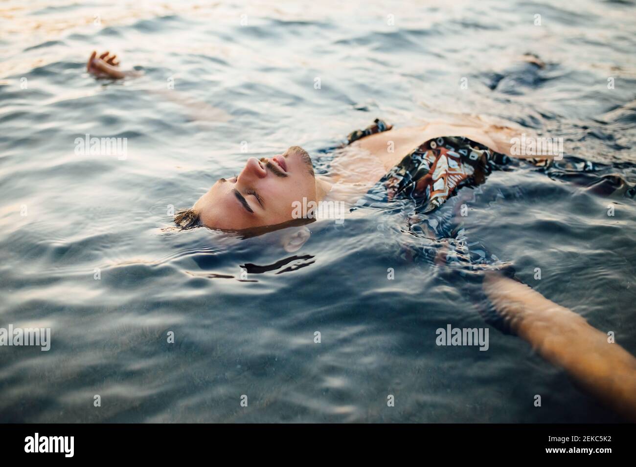 Junger Mann mit geschlossenen Augen trägt ein Hemd ohne Knöpfe, während er schwebt In Wasser Stockfoto