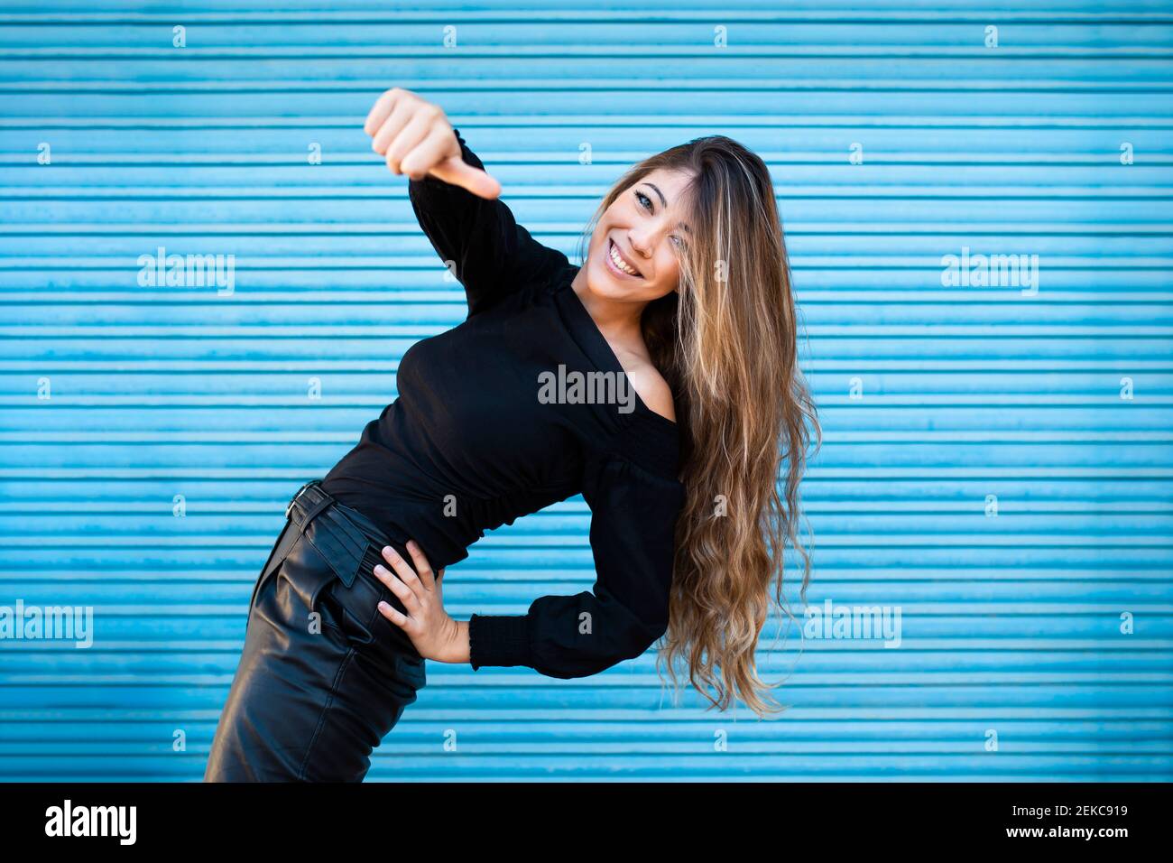 Junge Frau mit langen Haaren zeigt Daumen nach oben während stehen Gegen blaue Wand Stockfoto