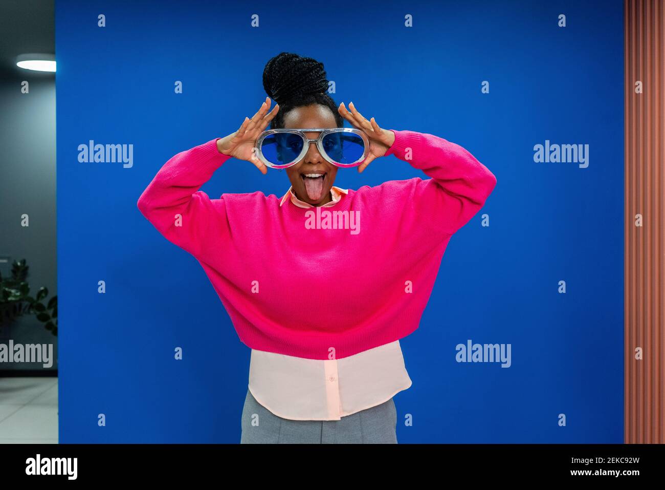 Junge Geschäftsfrau in überdimensionaler Sonnenbrille, die Zunge gegen Blau ausragt Wand am Arbeitsplatz Stockfoto