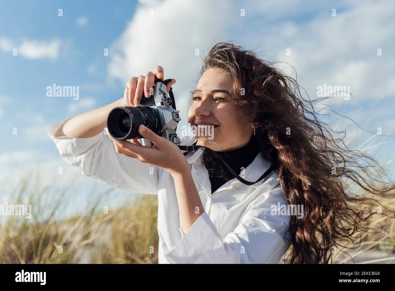 Lächelnde junge Frau, die am Strand durch die Vintage-Kamera fotografiert wolkiger Himmel Stockfoto Lächelnde junge Frau, die am Strand durch die Vintage-Kamera fotografiert wolkiger Himmel Stockfoto