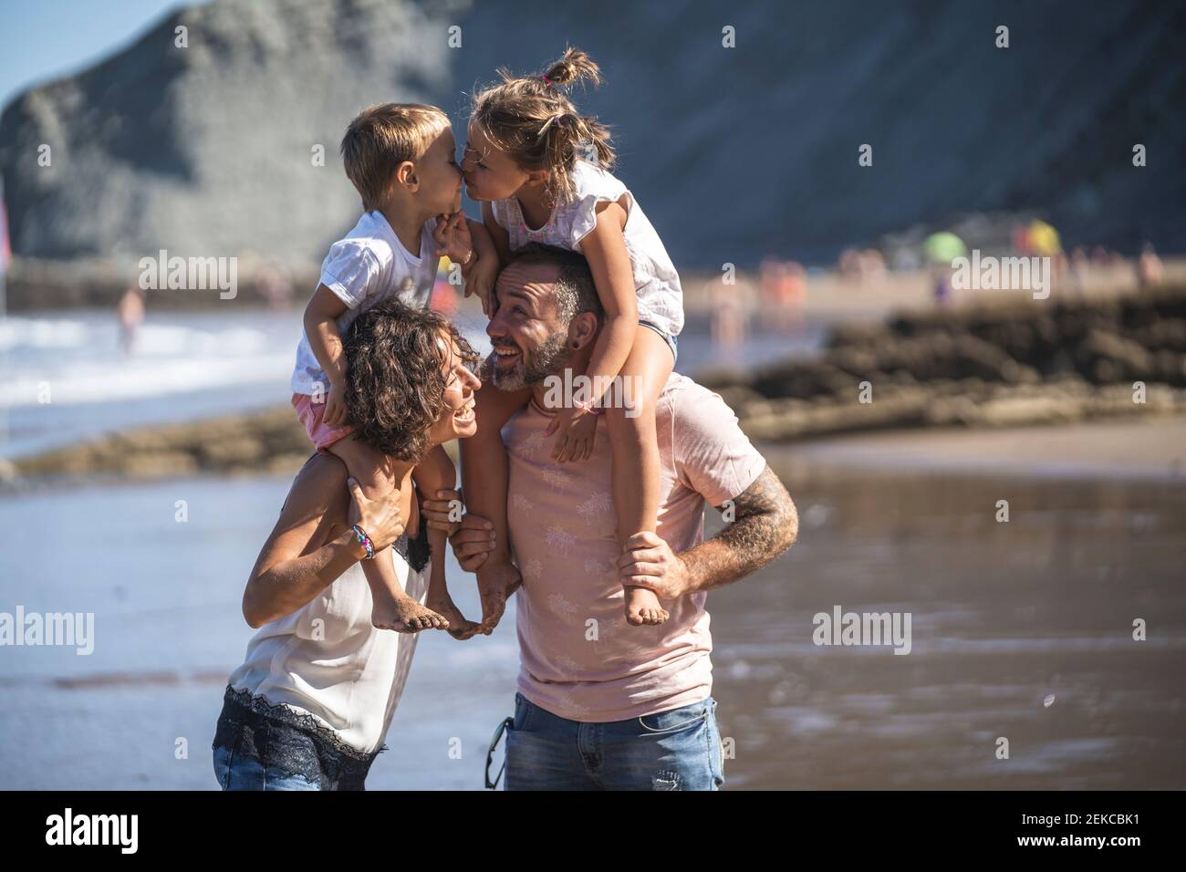 Geschwister küssen, während sie am Strand auf der Schulter der Eltern sitzen Stockfoto